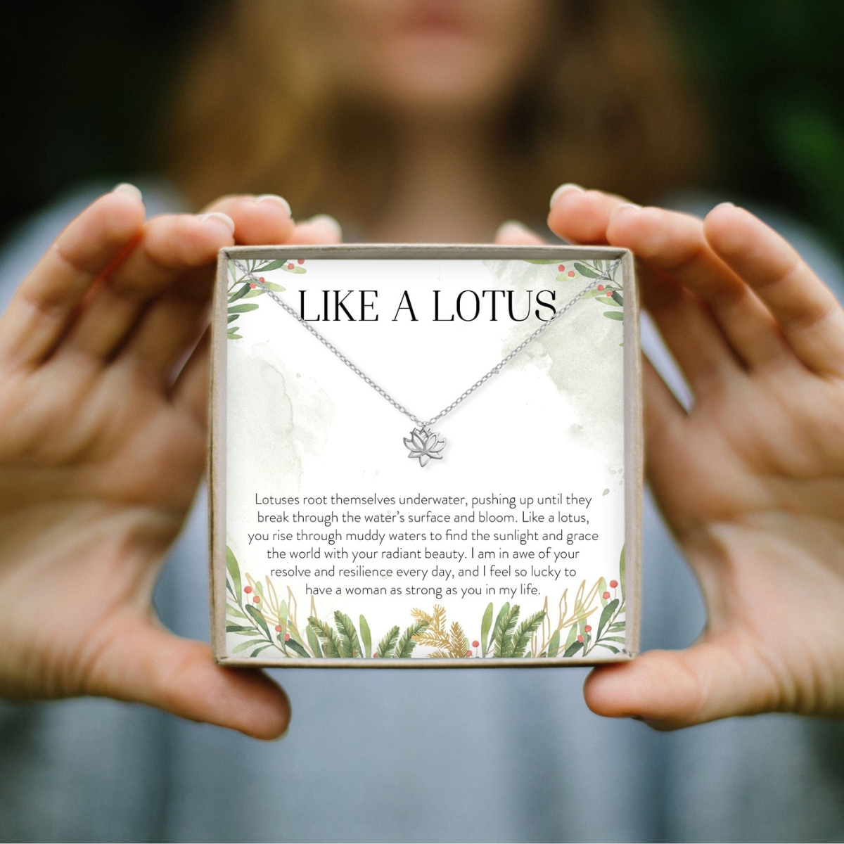 A person holds a square gift box containing a silver lotus necklace with a flower pendant, displayed against a softly blurred outdoor background. The front of the box reads “LIKE A LOTUS” and includes a sentimental message about lotus flowers.