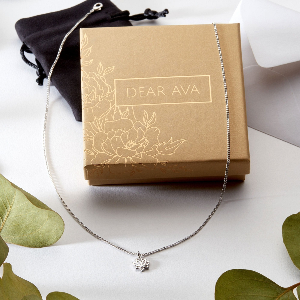 A silver chain necklace with a small flower-shaped pendant is draped over a brown gift box labeled 'DEAR AVA,' with a black drawstring pouch behind it on a white surface with green leaves around the edges.