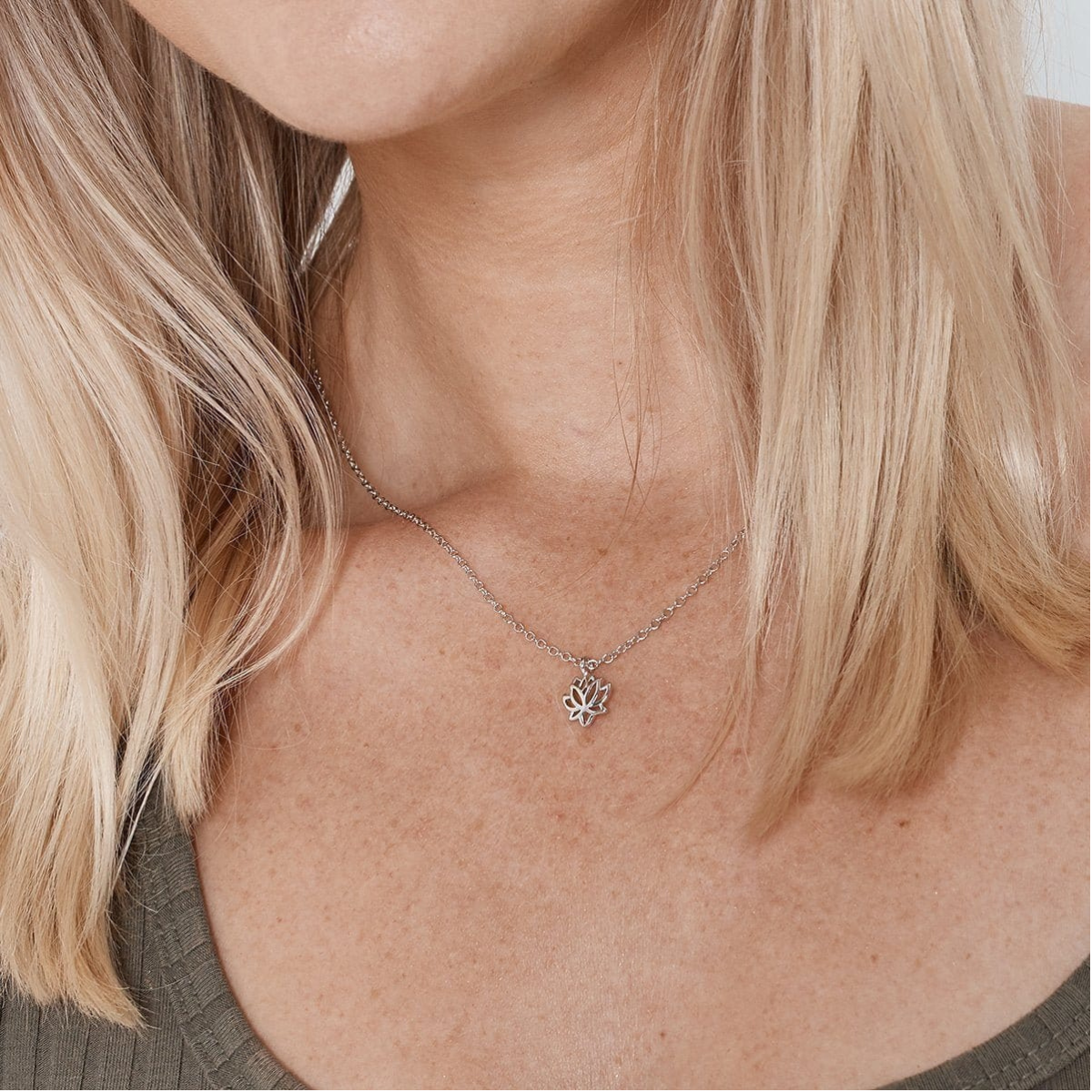 A close-up of a model's neck and chest wearing a delicate silver chain necklace with a small silver lotus flower pendant.